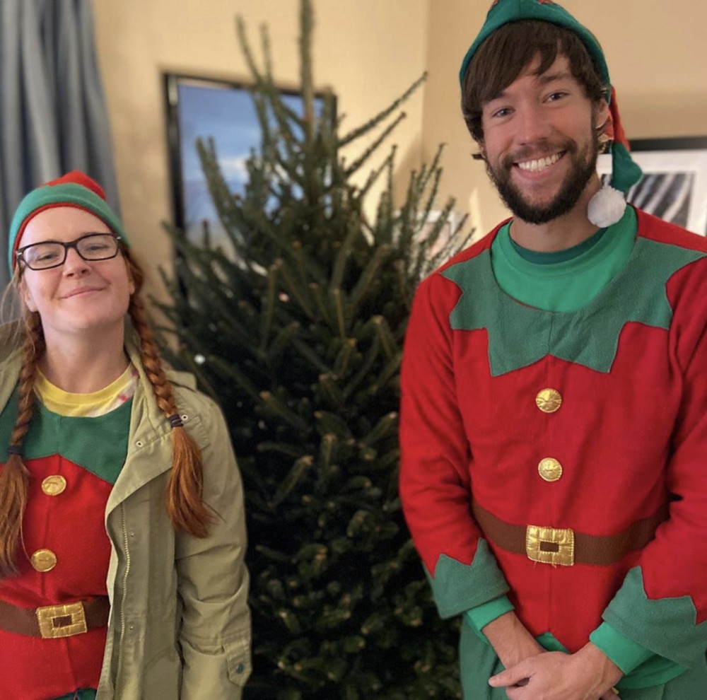 Two elves grinning beside a Fraser fir