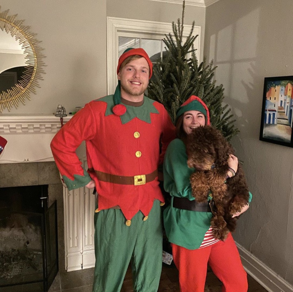 Two elves and a curly-haired dog beside a Fraser fir near a fireplace