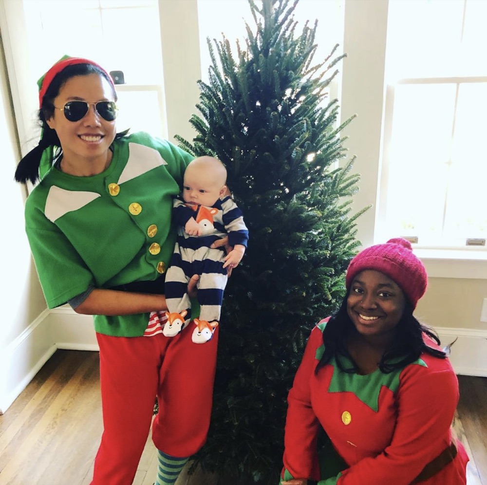 An elf holding a baby beside a Fraser fir by bright windows